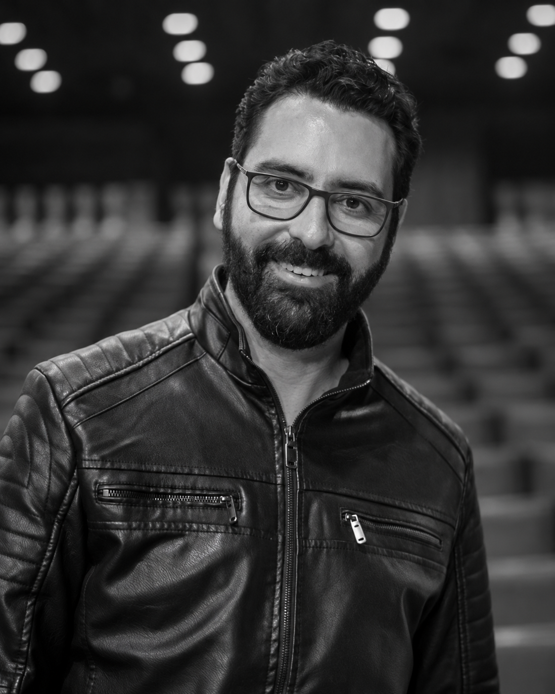 Retrato em preto e branco de Caio Sbierski, de óculos e jaqueta de couro, sorrindo.